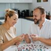 Dwie osoby siedzą przy stole w jasnej kuchni, układając puzzle. Na stole leży wiele kolorowych elementów puzzli.