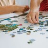 Na zdjęciu widać ręce układające puzzle na białym stole, z widocznymi fragmentami obrazka przedstawiającego krajobraz z jeziorem i drzewami. Puzzle są w różnych kolorach, a niektóre elementy są już połączone.