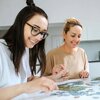 Na zdjęciu widać dwie osoby układające puzzle na stole w jasnym pomieszczeniu. Osoby są uśmiechnięte i skupione na układaniu elementów.