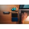 Na drewnianym blacie znajduje się niebiesko-czarna mysz komputerowa, obok której leży mały odbiornik USB oraz pendrive. Obok nich widoczny jest srebrny hub z logo "Verbatim", dwa zewnętrzne dyski twarde o fakturze przypominającej włókno węglowe oraz lapto