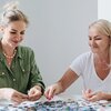 Na zdjęciu widać dwie osoby układające puzzle na białym stole. Na blacie rozłożone są różnokolorowe elementy puzzli.
