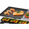 Na zdjęciu widoczny jest grill elektryczny z płytą grzewczą, na której znajdują się trzy krewetki i kawałek łososia z ziołami. Pod płytą znajdują się małe patelnie z roztopionym serem, plasterkami pomidora i liśćmi bazylii.