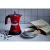 Czerwona kawiarka z napisem "Moka aroma" i "Ariete" stoi na blacie obok filiżanki z kawą i talerzyka z ciastkiem, a w tle leży otwarta książka. Na blacie rozsypane są ziarna kawy.
