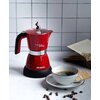 Czerwona kawiarka z napisem "Moka aroma" i "Ariete" stoi na blacie obok filiżanki z kawą i otwartej książki. Na blacie leżą również kilka ziaren kawy i dwa herbatniki.