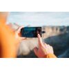 Na zdjęciu widać ręce trzymające smartfon z podłączonym pendrive'em, na ekranie widoczny jest interfejs aparatu fotograficznego. W tle rozciąga się rozmyty krajobraz górski.