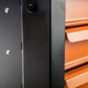 Zbliżenie na fragment szafki narzędziowej z czarną, matową powierzchnią i pomarańczowymi szufladami. Widoczne są detale konstrukcyjne, takie jak śruby mocujące.