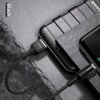 Czarny kabel USB - USB-C marki USAMS jest podłączony do urządzenia elektronicznego, które leży na ciemnej, teksturowanej powierzchni. Na zdjęciu widoczny jest napis "Black" oraz logo "USAMS" na wtyczce kabla.
