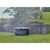 Na zdjęciu widoczny jest garaż dla robotów koszących GARDENA Sileno Minimo City i Life, umieszczony na trawniku, podczas deszczu. W tle znajduje się kamienna ściana oraz rośliny ozdobne.