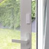 Na zdjęciu widoczny jest biały czujnik z napisem "NETATMO" zamontowany na framudze drzwi balkonowych, obok klamki. W tle znajduje się ogród z zielonym trawnikiem i roślinnością.