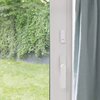 Na zdjęciu widoczny jest biały czujnik zamontowany na framudze drzwi balkonowych, obok uchylonej zasłony. W tle znajduje się ogród z zielonym trawnikiem i ścianą porośniętą bluszczem.