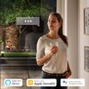 Kobieta stoi przy oknie, trzymając szklankę, a za nią widoczny jest ogród z drzewem i stosami drewna. Na zdjęciu znajdują się logotypy: "Works with Alexa", "Works with Apple HomeKit" oraz "Works with the Google Assistant".