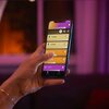 Na zdjęciu widać dłoń trzymającą smartfon z aplikacją do sterowania oświetleniem, na ekranie widoczne są różnokolorowe paski z nazwami: "Hue Signe", "Downlight", "Twilight" i "Flourish". W tle znajduje się rozmyte, ciepłe oświetlenie wnętrza.