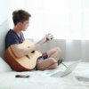 Na zdjęciu widać osobę siedzącą na łóżku z gitarą akustyczną, obok której leży otwarty laptop, myszka i smartfon. Kabel jest podłączony do gitary i laptopa, a w tle widoczne są jasne zasłony.