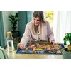 Na zdjęciu znajduje się osoba układająca puzzle przedstawiające postacie z bajki Disney "Królewna Śnieżka". Na stole obok puzzli stoi szklana karafka z korkiem oraz zielona roślina w doniczce.