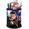 Na zdjęciu znajduje się przybornik w formie puzzli 3D z motywem z serii "Harry Potter", przedstawiający trzech młodych czarodziejów w strojach szkolnych z różdżkami. W przyborniku umieszczone są różne przybory, takie jak nożyczki, pędzle i ołówki.