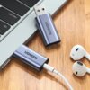 Na zdjęciu widoczny jest adapter UGREEN CM383 USB-A - Jack 3.5 mm podłączony do laptopa oraz para białych słuchawek z oznaczeniami "R" i "L" na drewnianej powierzchni. Adapter ma metaliczną obudowę z napisem "UGREEN".