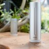 Na zdjęciu widoczny jest cylindryczny, srebrny moduł z napisem "Netatmo" umieszczony na drewnianej powierzchni, z rozmytym tłem przedstawiającym roślinność. W tle znajduje się drugi, podobny moduł.