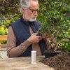 Na zdjęciu widoczny jest biały, cylindryczny produkt z napisem "Netatmo" umieszczony na drewnianym stole obok doniczki z bonsai. W tle znajduje się osoba przycinająca drzewko, a otoczenie jest zielone i pełne roślinności.