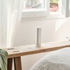 Na zdjęciu znajduje się cylindryczne urządzenie stojące na drewnianej ławce, z widocznym logo "Netatmo" na dole. W tle widać otwarte okno z białą ramą i zasłoną, przez które wpada naturalne światło.