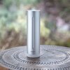Srebrny, cylindryczny produkt z napisem "Netatmo" na dole, stoi na drewnianym, ozdobnym stole. Tło jest rozmyte, z zielonymi i brązowymi odcieniami.
