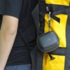 Czarne etui na głośnik GRAVASTAR do Venus jest przymocowane karabińczykiem do żółto-czarnego plecaka. Na etui widoczne jest logo producenta.