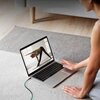Osoba siedzi na dywanie przed otwartym laptopem, na ekranie którego wyświetlany jest obraz osoby wykonującej ćwiczenia rozciągające. Laptop jest podłączony do zasilania za pomocą czarnego kabla USB-C.