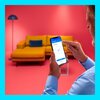 Na zdjęciu widać osobę trzymającą smartfon z aplikacją, na której ekranie widnieje napis "Smart pairing" oraz "Pairing completed" z dużym niebieskim znakiem potwierdzenia. W tle znajduje się żółta sofa z niebieską poduszką oraz lampa stojąca, a całość utr