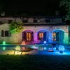 Na zdjęciu widać kamienny dom z łukowatymi oknami i drzwiami, ozdobiony kolorowymi lampkami LED, które tworzą różnobarwne iluminacje na elewacji i wokół basenu. W ogrodzie znajdują się rośliny oraz leżaki, a całość jest oświetlona w różnych odcieniach, tw