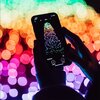 Na zdjęciu widać smartfon z aplikacją wyświetlającą kolorowe światełka ułożone w kształt choinki, na tle rozmytych, wielobarwnych świateł. Na ekranie telefonu widoczny jest napis "Rainbow" oraz ikony z napisami "MODYF." i "APLIKUJ".