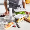Na zdjęciu znajduje się szklany pojemnik ZWILLING Fresh & Save o pojemności 0,35 L, obok którego stoi biała pompka próżniowa z logo ZWILLING. W tle widoczne są inne pojemniki z jedzeniem, miska z wiśniami oraz deska z cytrynami i nożem.