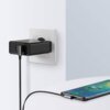 Na zdjęciu widać czarną ładowarkę podłączoną do gniazdka elektrycznego, z której wychodzi szary kabel USB-C podłączony do smartfona leżącego na powierzchni. Na ekranie telefonu widoczny jest symbol ładowania z procentowym wskaźnikiem naładowania baterii.