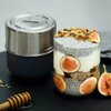 Na zdjęciu znajduje się metalowy lunch box z logo "BLACK+BLUM" oraz szklanka z warstwami chia, jogurtu, granoli i plasterkami fig. Obok leży drewniana łyżka do miodu i świeża figa.