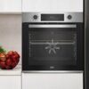 Na zdjęciu znajduje się piekarnik elektryczny w zabudowie, wykonany ze stali nierdzewnej, z wyświetlaczem pokazującym "00:34" oraz dwoma pokrętłami. Na blacie obok piekarnika stoi miska z czerwonymi jabłkami, a na froncie piekarnika widoczne jest logo "be