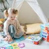 Na zdjęciu widać dziecko bawiące się kartami z gry edukacyjnej "TREFL ABC malucha Farma", obok znajduje się stos pudełek z grami, na których widoczne są napisy: "Pory roku", "Zwierzak", "Przeciwieństwa", "Pojazdy", "Liczby i kształty". W tle znajduje się 