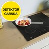 Na zdjęciu znajduje się czarna płyta indukcyjna z białą patelnią, na której smażone są kawałki jedzenia na czerwonym tle. W lewym górnym rogu widoczny jest żółty okrąg z napisem "DETEKTOR GARNKA".