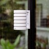 Biała osłona stacji pogodowej zamontowana na ciemnej powierzchni, z widocznym logo "Netatmo" na dolnej części. W tle rozmyte zielone liście i elementy architektury.
