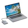 Na zdjęciu znajduje się otwarty laptop z wyświetlaczem pokazującym plażę z palmami, do którego podłączony jest zewnętrzny dysk twardy z widocznym logo "LACIE". Dysk jest połączony z laptopem za pomocą kabla USB.
