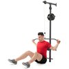 Na zdjęciu znajduje się wyciąg do ćwiczeń zamontowany na ścianie, z widocznym logo "SmartGym" na górze. Mężczyzna w czerwonej koszulce i czarnych spodenkach siedzi na podłodze, trzymając drążek wyciągu.