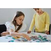 Dwoje dzieci układa puzzle na okrągłym stole, skupiając się na dopasowywaniu elementów. Na stole widoczne są różnokolorowe kawałki puzzli oraz fragment obrazka z pieskami.