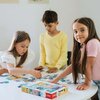 Na zdjęciu widać troje dzieci układających puzzle na białym stole, obok znajduje się pudełko z puzzlami "Puzzle TREFL W świecie piesków (100 elementów)". Na pudełku widoczne są kolorowe ilustracje piesków oraz logo "Trefl".