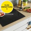 Na zdjęciu znajduje się czarna płyta indukcyjna zamontowana na blacie kuchennym, na której stoi patelnia z jedzeniem. Obok płyty widoczny jest żółty okrąg z napisem "KLASA ENERGETYCZNA A", a na blacie znajdują się różne produkty spożywcze i przyprawy.