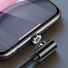Na zdjęciu widać fragment smartfona z podłączonym magnetycznym kablem USB, który ma czarną, plecioną osłonę. Kabel jest połączony z telefonem za pomocą okrągłego, metalowego złącza, a na ekranie urządzenia widoczny jest biały pasek.