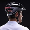 Na zdjęciu widoczny jest szaro-czarny kask rowerowy z lampką, z napisem "LIVALL" na boku. Kask ma wbudowane czerwone diody LED z tyłu, a osoba go nosząca ma na sobie białą koszulkę.