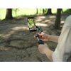 Osoba trzyma gimbal z zamocowanym smartfonem, na ekranie którego widać nagrywany obraz zielonego krajobrazu z drzewami. Tło zdjęcia jest rozmyte, ukazując parkową scenerię z drzewami i cieniem na ziemi.