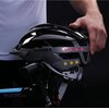 Szaro-czarny kask rowerowy z lampką, oznaczony napisem "LIVALL", jest trzymany nad kierownicą roweru. Na kasku widoczne są świecące czerwone i żółte diody.