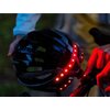 Czarny kask rowerowy z wbudowanymi czerwonymi diodami LED jest trzymany w rękach, a obok znajduje się ręka naciskająca przycisk na kierownicy roweru. Kask ma nowoczesny design z otworami wentylacyjnymi.