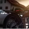 Na zdjęciu widoczny jest czarny kask z wbudowaną lampką, umieszczony na rowerze, z wyraźnie widocznym czerwonym światłem. W tle znajduje się rozmyty obraz budynku i samochodu.