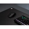 Na zdjęciu znajduje się czarna mysz komputerowa na dużej podkładce z widocznym logo Corsair. Obok myszy widać fragment klawiatury z podświetlanymi klawiszami w różnych kolorach.