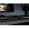Na zdjęciu znajduje się czarna mysz komputerowa z podświetleniem RGB, umieszczona na ciemnej podkładce. W tle widoczny jest monitor z abstrakcyjną grafiką oraz stojak na słuchawki z podświetleniem.