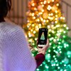 Osoba trzyma smartfon z aplikacją wyświetlającą choinkę z kolorowymi światłami, a w tle widać rozmyte, świecące lampki choinkowe w różnych kolorach.